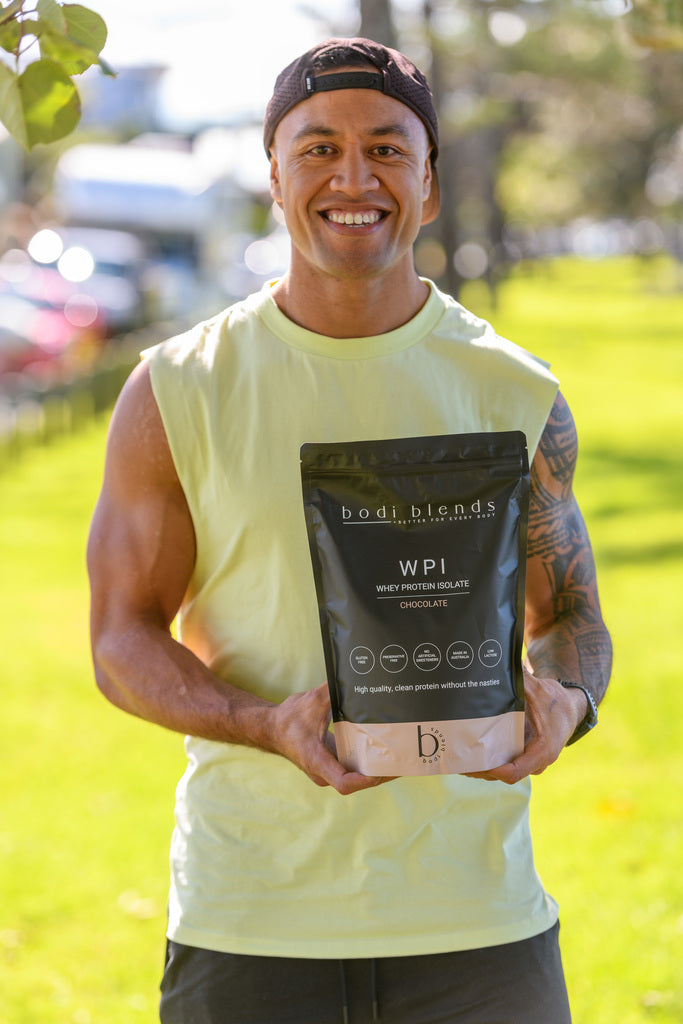 A man in a yellow singlet and a backwards black cap smiling holding a black and brown stand up pouch of chocolate flavoured whey protein powder