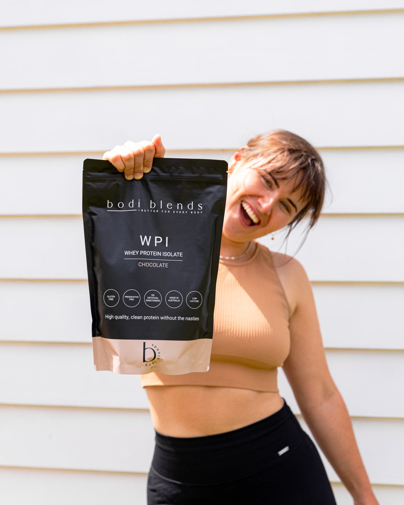 A girl in a brown crop top and black tights smiling holding out a black and brown stand up pouch of chocolate flavoured whey protein powder.