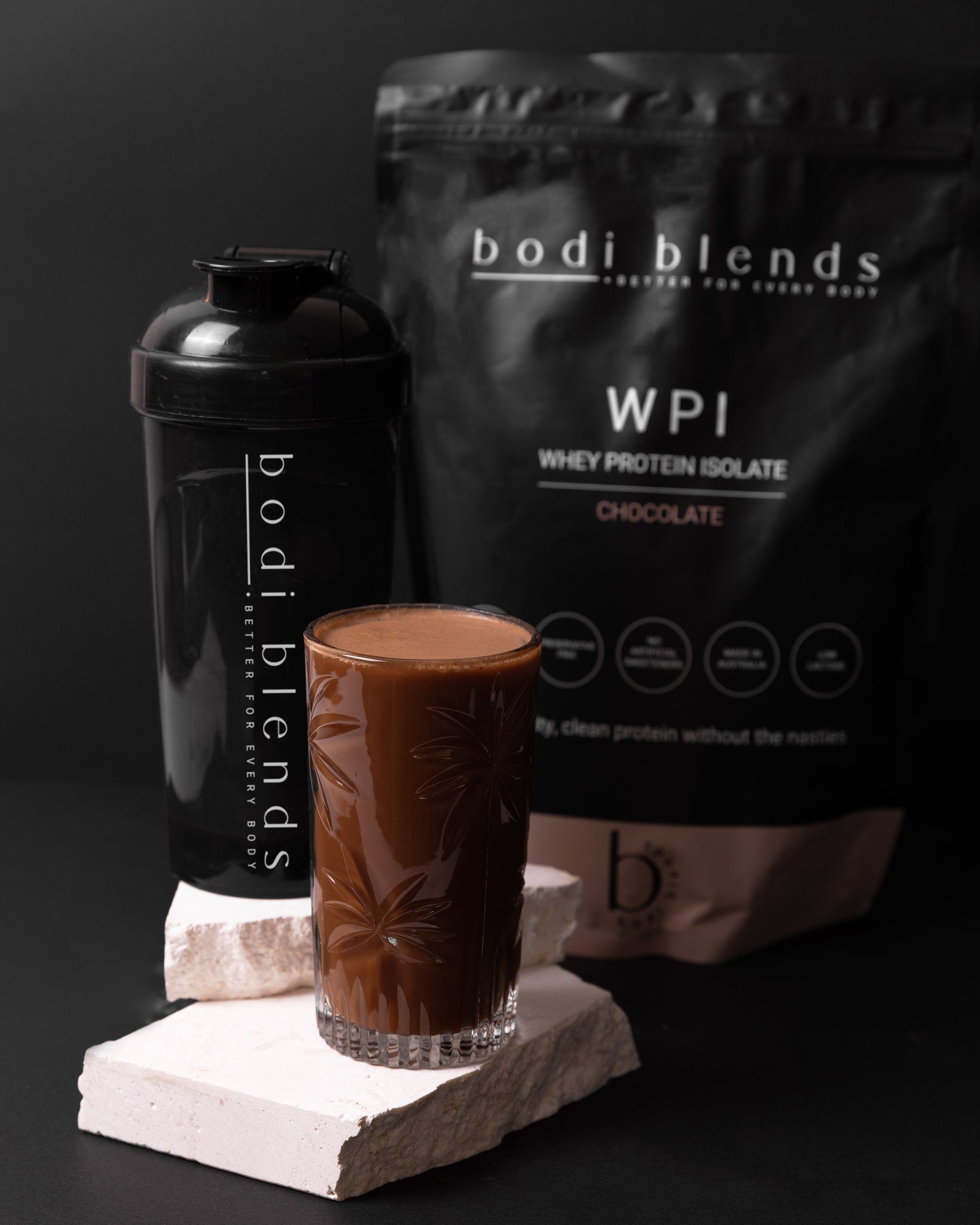 A glass sitting on a piece of rock with brown liquid inside. A black protein shaker sitting on a rock behind the glass with a white Bodi Blends logo down the side. A black and brown stand up pouch behind the shaker and glass with brand logo and nutritional information on it.
